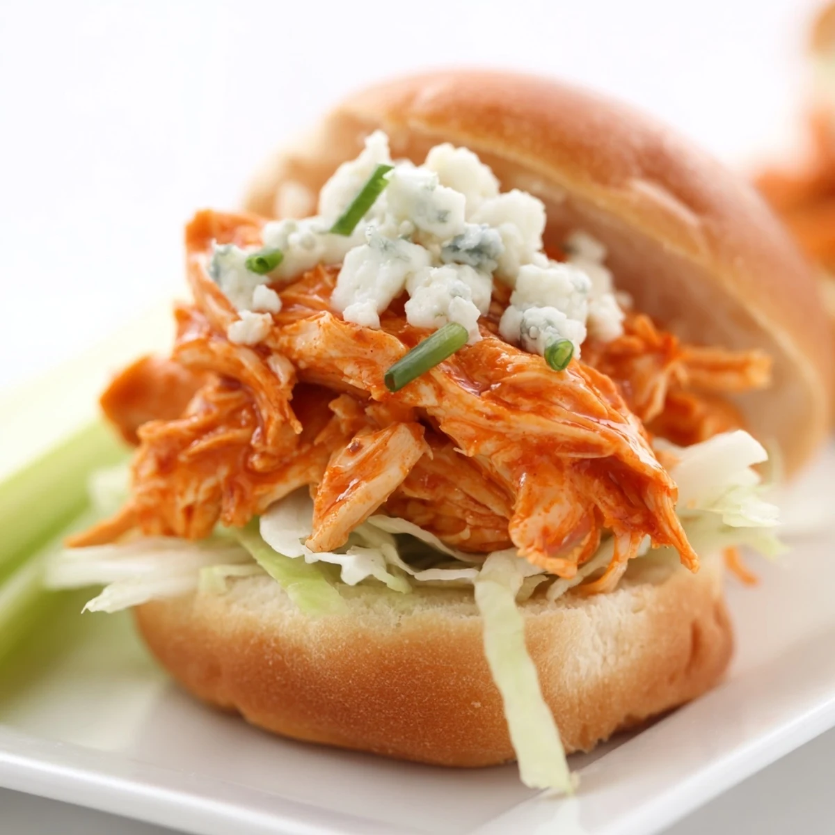 Freshly assembled Buffalo chicken sliders featuring tender meat, crunchy celery and red onion, blue cheese crumbles, and soft buns on a wooden serving board.  
