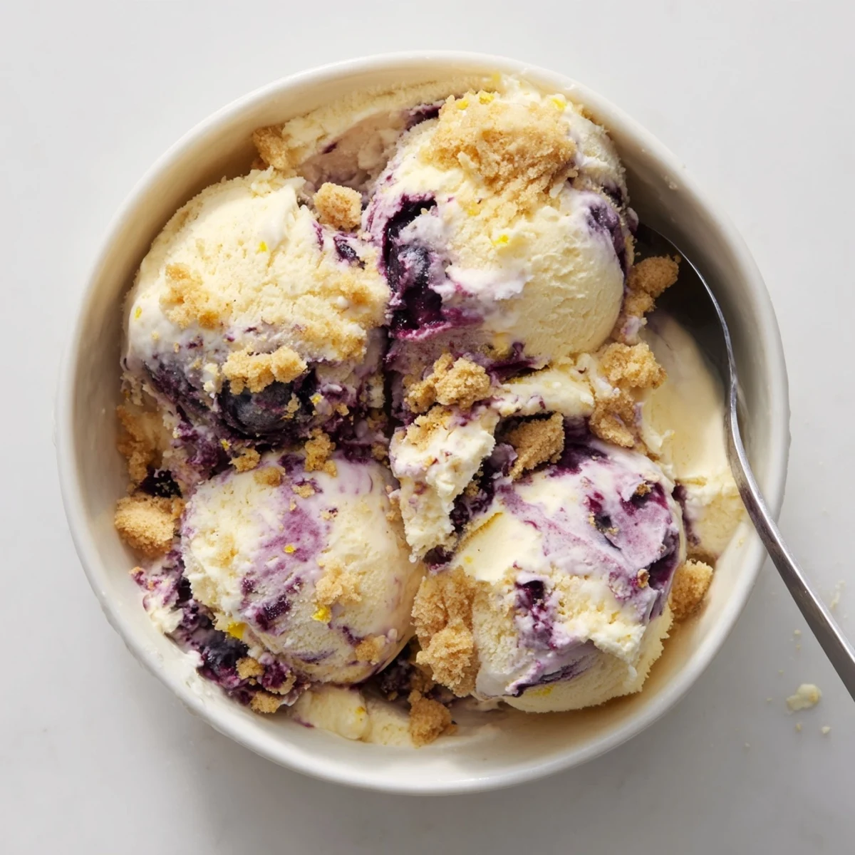 Scoop of Lemon Blueberry Cheesecake Ice Cream in a glass bowl, showing creamy texture and blueberry swirls, served with a graham cracker crust crumble on the side.