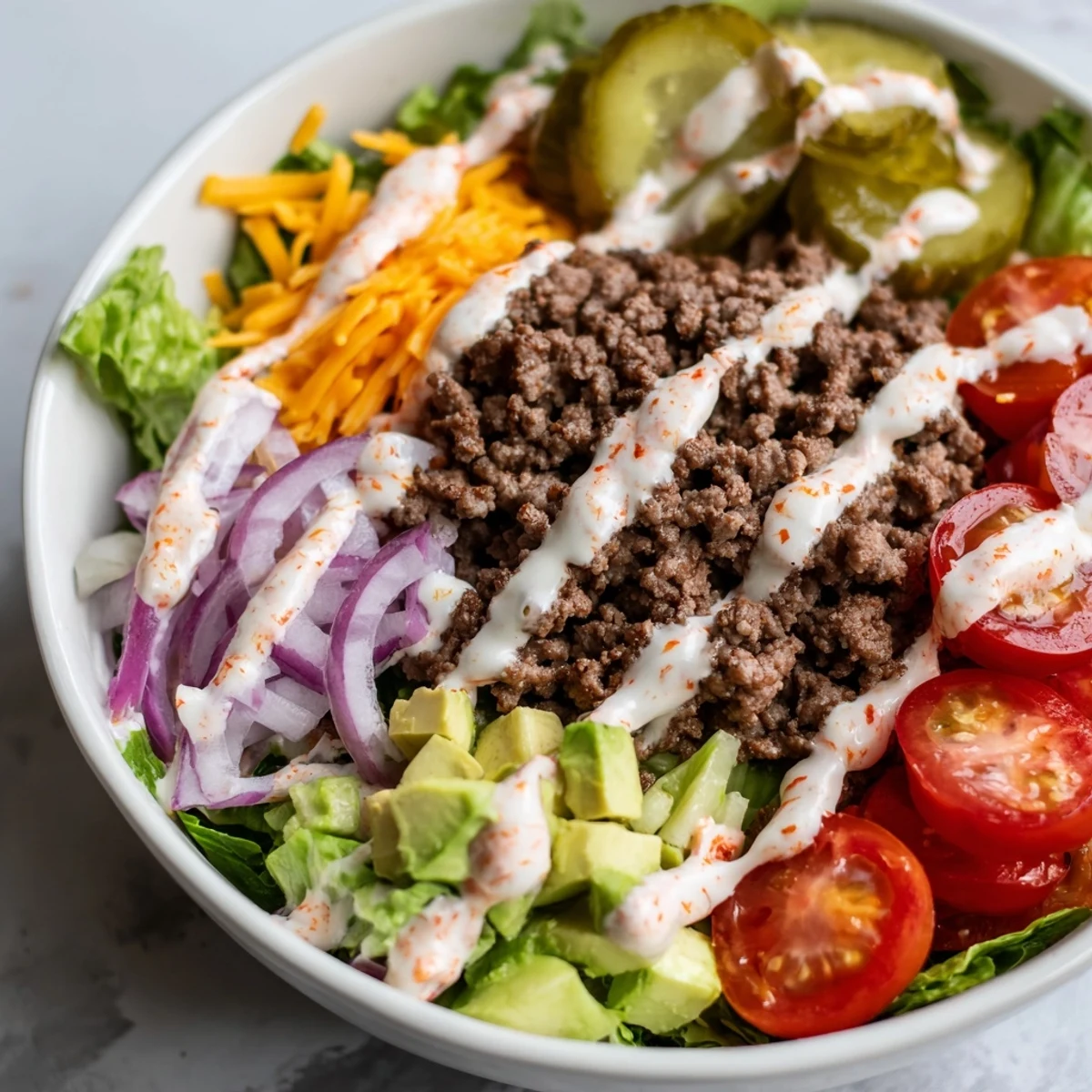 Fresh low-carb burger bowl featuring juicy seasoned beef, vibrant red onion, ripe avocado chunks, zesty pickle slices, and tangy homemade burger sauce drizzled on top.