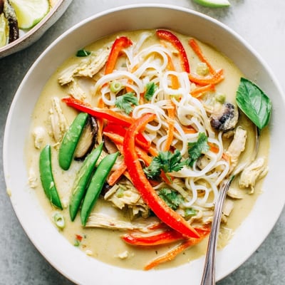 Serving bowls are filled with steaming Thai Green Curry Chicken Noodle Soup, featuring creamy coconut broth, colorful veggies, and a sprinkle of red chili.