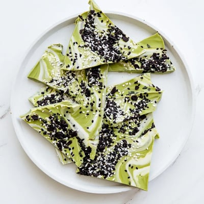 Cracked shards of Black Sesame Matcha Bark on a marble board, featuring a marbled green and white surface with black sesame seeds and a hint of flaky salt. 