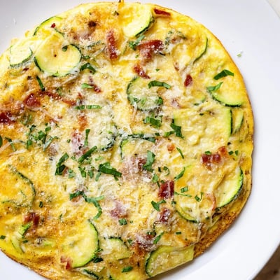 A close-up view of Zucchini Herb and Pancetta Frittata featuring herbs, melted Parmesan, and a rustic oven-baked finish on a skillet.
